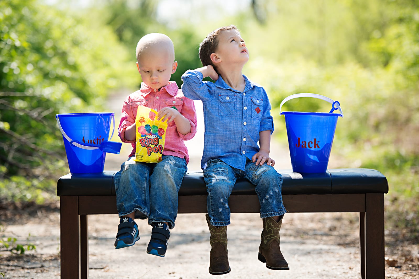 Two young boys sit side by side on a bench along a sunny path, a bright blue sand pail on the bench beside each of them; the boy on the left has no hair and holds a bag of ring-shaped candy, and the boy on the right wears a denim shirt and cowboy boots.