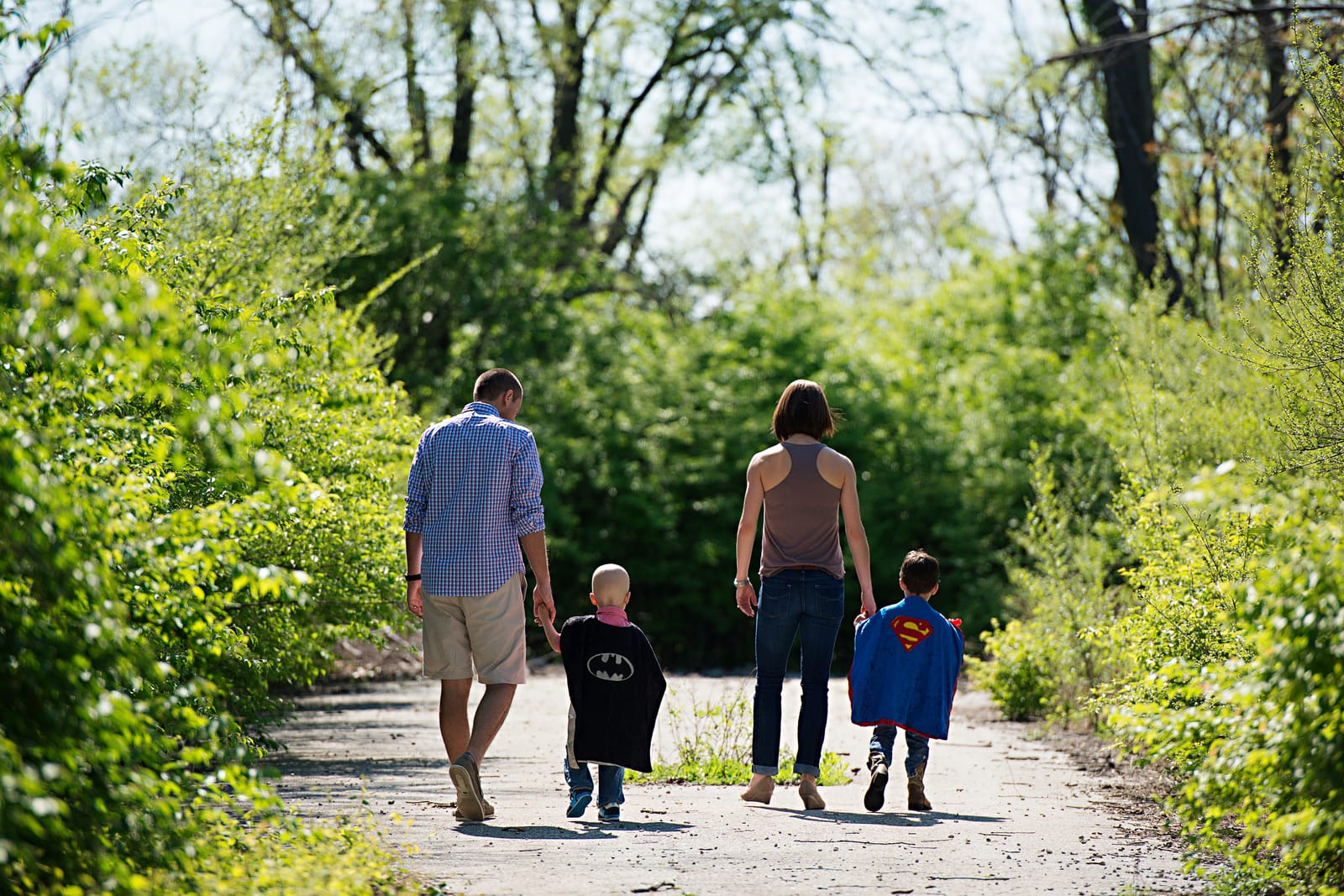 A family of four seen from behind walks down a sunlit, tree-lined path holding hands, the two young children wearing superhero capes — one a black Batman cape, the other a red-and-blue Superman cape.