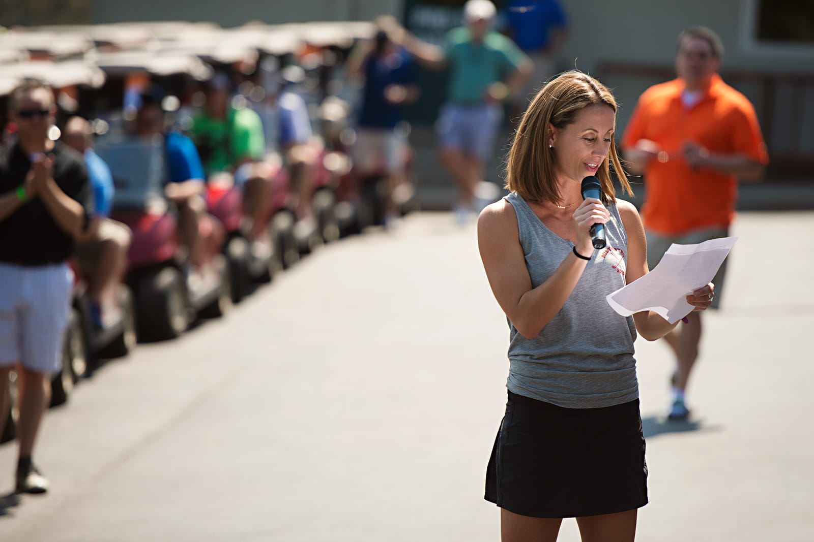 A woman speaks into a handheld microphone and reads from a sheet of paper at a golf tournament, with a long line of golf carts and participants lined up behind her on a sunny day.