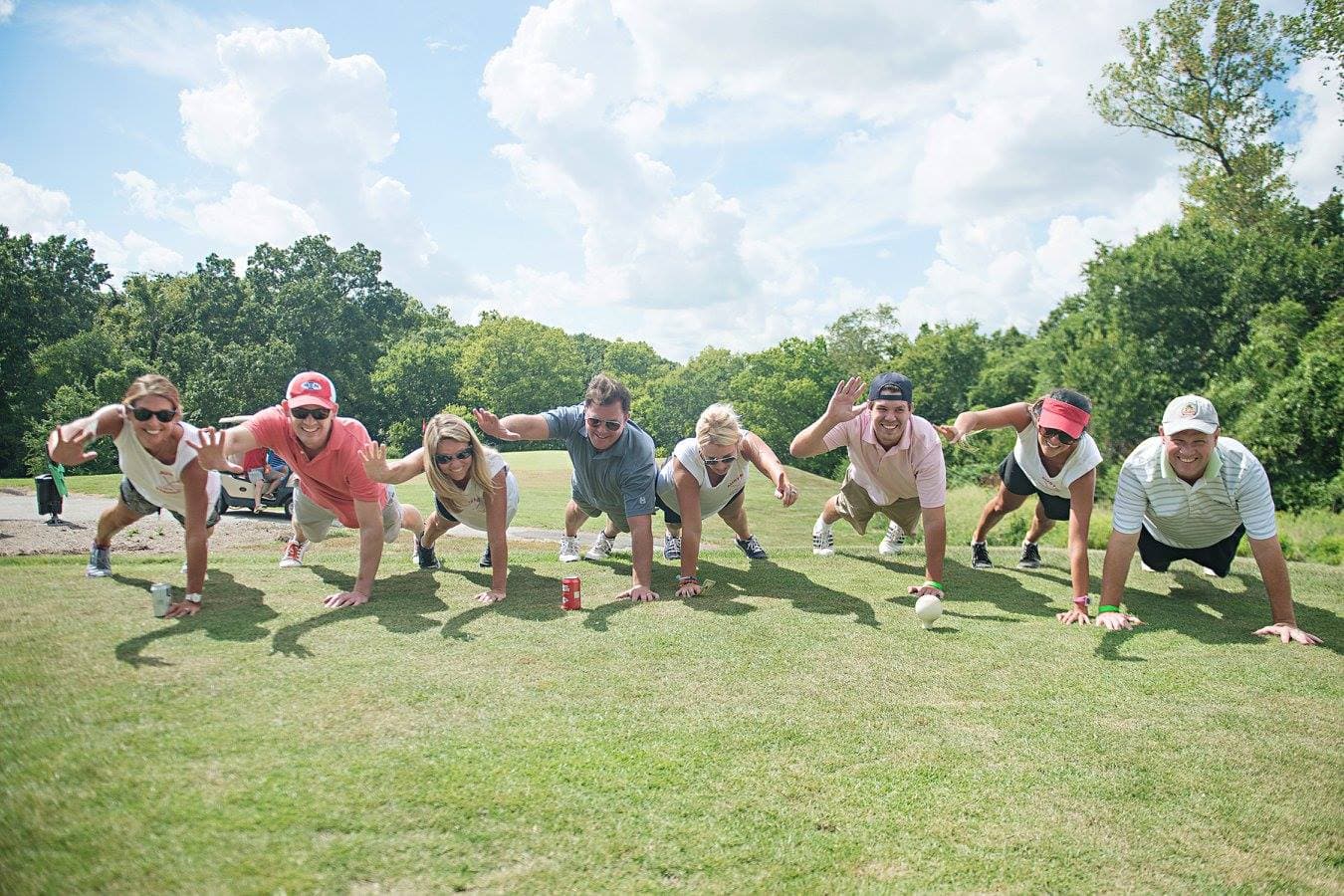Eight people in golf attire do push-ups in a row on a golf course fairway, hands on the grass and smiling toward the camera, under a bright sky with scattered clouds.